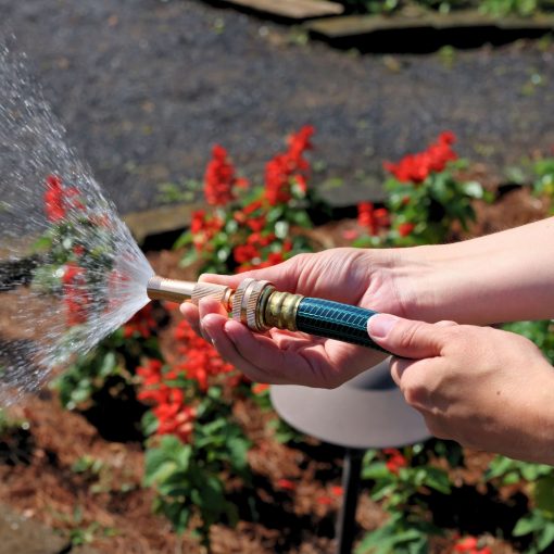 A person's hands holding a garden hose with a nozzle, spraying water on red flowers in a sunlit garden with foliage in the background.