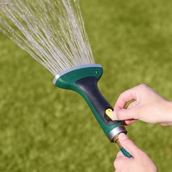 A person's hands are holding a green and black garden hose nozzle, spraying a fan-shaped water pattern onto a lush green lawn in daylight.