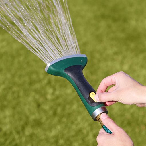 A person's hands are holding a green and black garden hose nozzle, spraying a fan-shaped water pattern onto a lush green lawn in daylight.