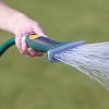 This image shows a close-up of a person's hand holding a green and grey hose nozzle, spraying a fan-shaped pattern of water onto grass.