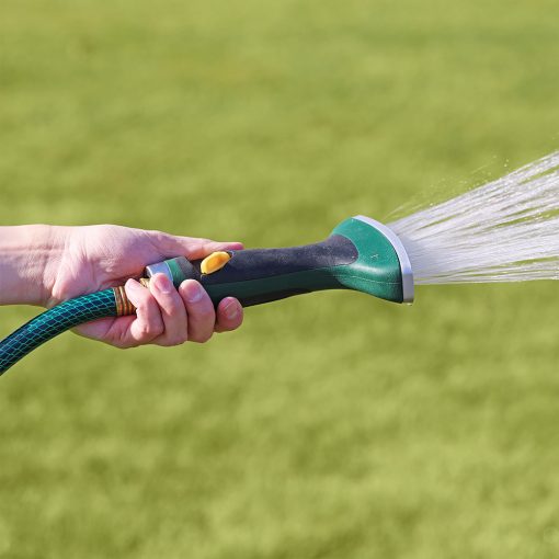 A person's hand is holding a green and black garden hose nozzle, spraying water onto a lush green lawn in bright daylight.