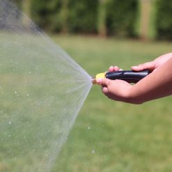 A person's hand holds a garden hose with a nozzle, spraying water forcefully in a wide arc. A green lawn and trees are visible in the background.