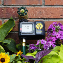 An electronic water timer is attached to a spigot on a brick wall, surrounded by lush green leaves and vibrant purple flowers in a garden setting.