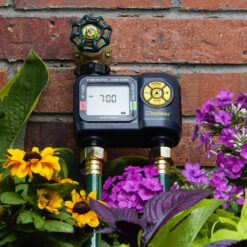 A digital water timer attached to a brick wall amidst colorful flowers, indicating a programmed irrigation system in a garden setting.