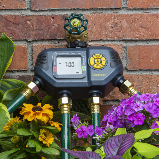 This image shows a digital watering timer with two outlets attached to a green hose bib against a brick wall, surrounded by vibrant flowers.