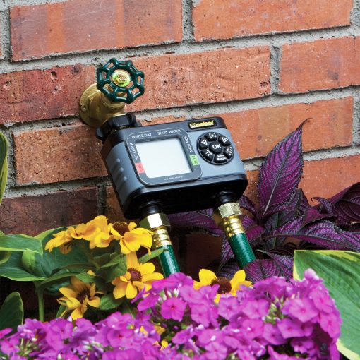 This is a digital water timer attached to a brass spigot on a red brick wall, surrounded by colorful flowers and green foliage.