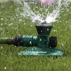 A green garden sprinkler is attached to a hose and is actively watering the grass. Water is spraying out in various directions against a sunny backdrop.