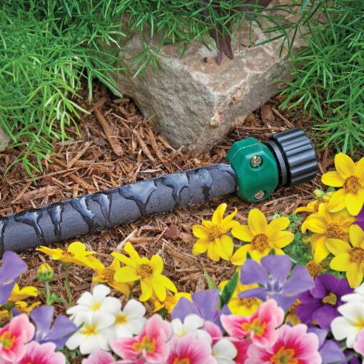 This image shows a soaker hose lying on the ground among colorful flowers, with mulch and a stone nearby, used for efficient garden watering.