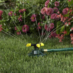 An oscillating sprinkler is attached to a green hose, watering a lush lawn with a background of vibrant pink and red flowers in a garden.