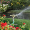 An oscillating sprinkler is watering a lush green lawn surrounded by colorful flowers, under a sunny sky with blurred background vegetation.