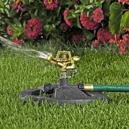 An oscillating sprinkler is attached to a green hose on lush green grass, spraying water, with vibrant pink and red flowers in the background.