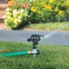 An oscillating lawn sprinkler is attached to a green garden hose, watering grass with a backdrop of colorful flowers and a brick-lined flowerbed.