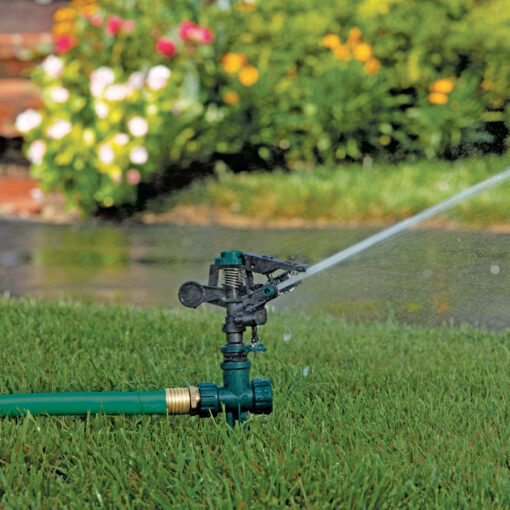 An oscillating sprinkler is attached to a green hose on a well-maintained lawn, spraying water. In the background, there are colorful flowers and shrubs.