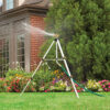 A tripod sprinkler is actively watering a lush garden with a variety of colorful flowers in front of a brick house with greenery.