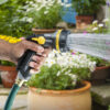 A person's hand is holding a hose with a spray nozzle, watering plants in a vibrant, flower-filled garden setting with various pots and greenery.