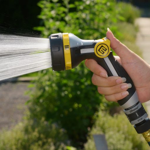 A person's hand is holding a garden hose nozzle, spraying a jet of water. The surrounding area is sunlit with greenery in the background.