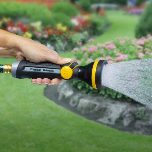 A person is holding a garden hose with a nozzle, spraying water on plants in a colorful blooming garden with a well-maintained lawn and flowers.