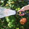 A person's hand is holding a garden hose with a nozzle, spraying water on plants in bright sunlight, with a blurred green background.