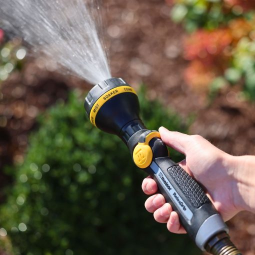 A person's hand is holding a black and yellow garden hose nozzle, spraying water, with plants and flowers softly focused in the background.