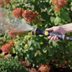 A person is holding a garden hose nozzle, spraying water onto lush plants with pinkish blooms, in bright, sunlit conditions, suggesting garden maintenance or watering.