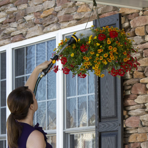 A person is watering a hanging basket of colorful flowers using a long nozzle attached to a hose outside a house with a stone facade.