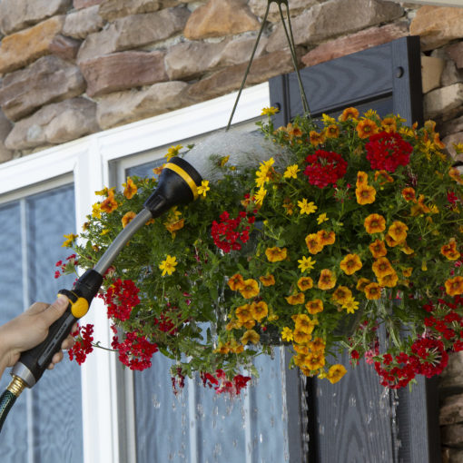 A person is watering a vibrant hanging flower basket with a hose, against a backdrop of a stone wall and window. Droplets are visible in the air.