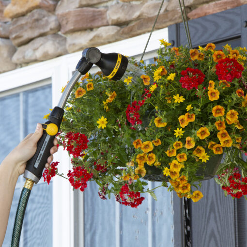 A person's hand is holding a hose with a spray nozzle, watering a vibrant hanging basket of red and yellow flowers in front of a house.