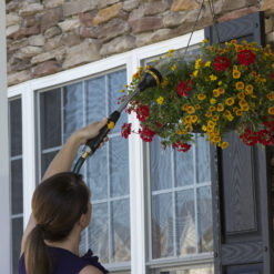 A person is watering a vibrant hanging flower basket with a hose nozzle, beside a door with windows, against a stone-clad house wall.