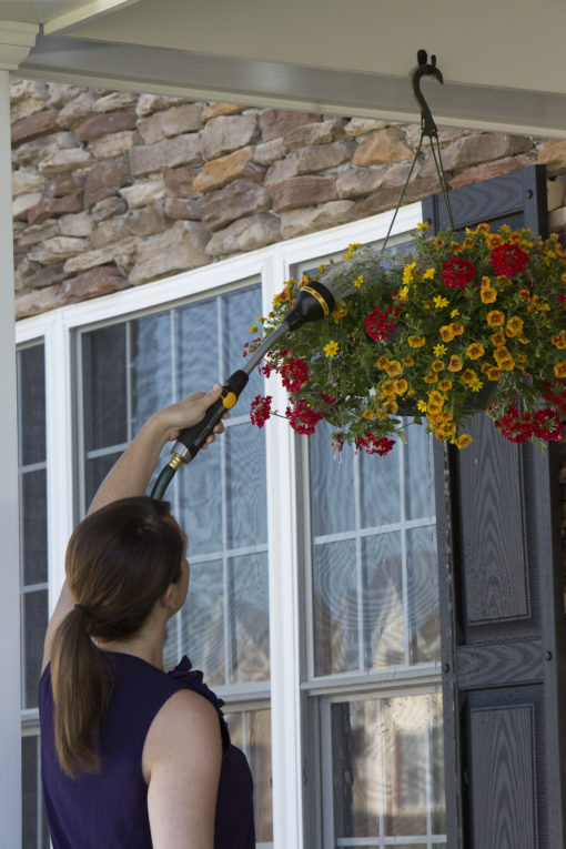 A person is watering a vibrant hanging flower basket with a hose nozzle, beside a door with windows, against a stone-clad house wall.