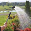 A person's hands hold a garden hose with a spray nozzle, watering plants. The background features lush greenery, a pond, and a clear sky.