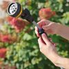 A person's hands are extending a watering wand amidst a blurry background of lush greenery with hints of pink and red flowers.