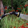 A garden hose with a spray nozzle is watering plants. Water jets are visible against a background of vibrant red flowers and green foliage.