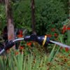 A garden hose with a spray nozzle attached emits a jet of water against a backdrop of lush greenery and red flowers, with a blurry person in the distance.