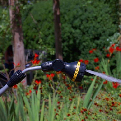 A garden hose with a spray nozzle attached emits a jet of water against a backdrop of lush greenery and red flowers, with a blurry person in the distance.