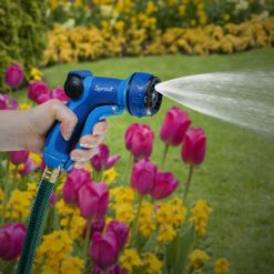 A person's hand is holding a blue garden hose nozzle, spraying water among pink and yellow flowers in a vibrant, well-maintained garden backdrop.