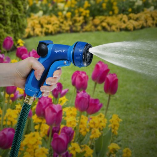 A person's hand is holding a blue garden hose nozzle, spraying water among pink and yellow flowers in a vibrant, well-maintained garden backdrop.