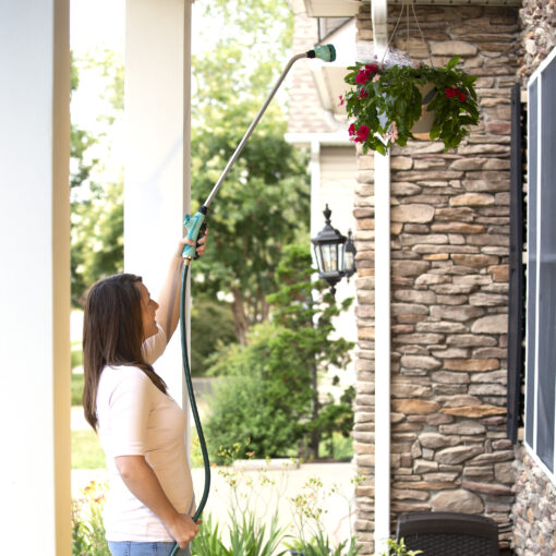 A person is using a long-handled watering wand to water a hanging plant with pink flowers outside a house with stone pillars.