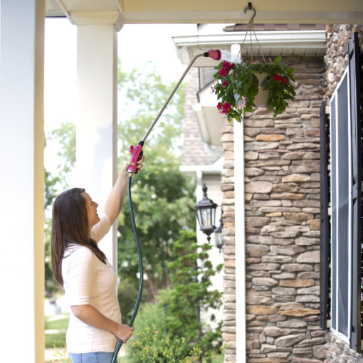 A person is watering a hanging flower basket with a hose in front of a house with stone accents. It's a sunny day with greenery around.