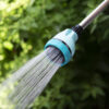 A garden hose nozzle is attached to a hose, spraying water with a gentle shower setting against a backdrop of lush green foliage.