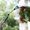 A person is watering hanging pink flowers using a garden hose with a spray nozzle, creating a mist, with a blurred background of a house.