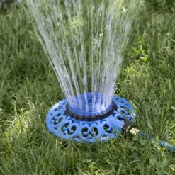 A circular blue lawn sprinkler is connected to a green hose, spraying water upwards onto green grass, providing irrigation under bright daylight conditions.