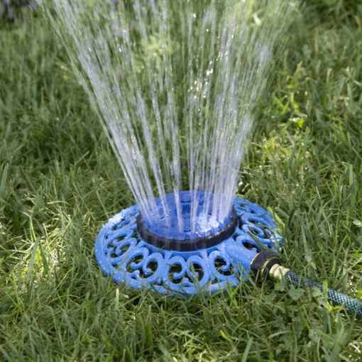 A circular blue lawn sprinkler is connected to a green hose, spraying water upwards onto green grass, providing irrigation under bright daylight conditions.