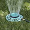 A circular lawn sprinkler is connected to a hose, spraying water upward in a fountain-like pattern onto the surrounding green grass under sunlight.