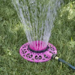 A pink circular lawn sprinkler connected to a hose is watering the grass, with water streams spraying upwards and outwards, soaking the green lawn.