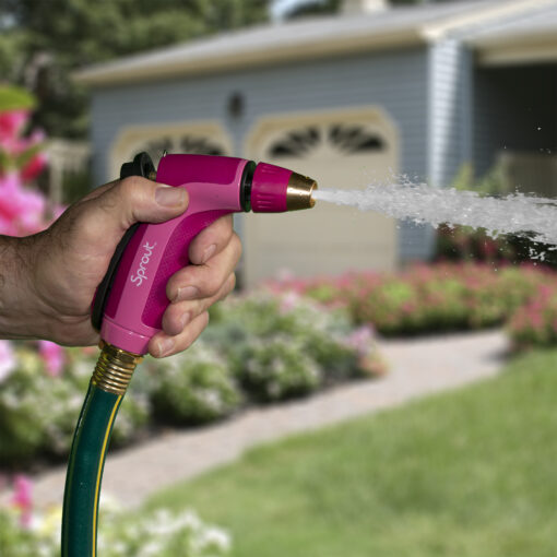 A person's hand is holding a pink garden hose nozzle, spraying water, with a blurred background of a house and flowering garden.