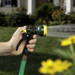 A person's hand is holding a green and yellow garden hose with a nozzle, spraying water, with a blurred background of a lawn and flowers.