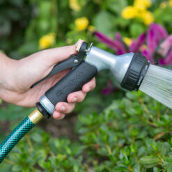 A person's hand is holding a black and grey garden hose nozzle, spraying water among vibrant green plants and colorful flowers in a garden.