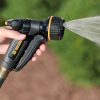 A person is holding a black and yellow garden hose nozzle, spraying water in a mist pattern, against a blurred green background outdoors.