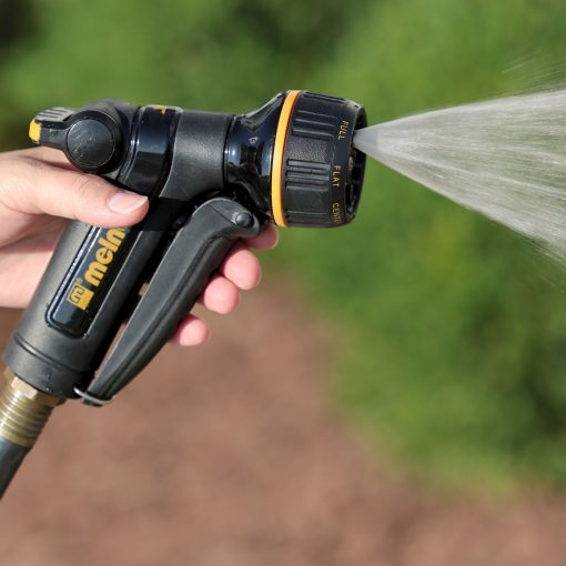 A person is holding a black and yellow garden hose nozzle, spraying water in a mist pattern, against a blurred green background outdoors.