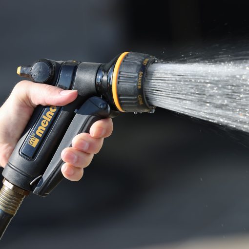 A person's hand is holding a black and orange garden hose nozzle, spraying water with a mist setting, against a blurred background.